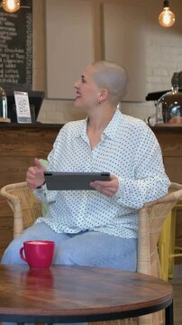 Vertical video: Receiving red mug, shaved woman in dotted blouse tapping tablet and sipping at cafe