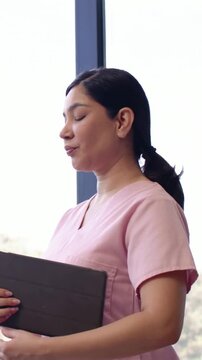 Vertical video: Glancing out then opening tablet, nurse in pink scrubs reviewing notes by mullion