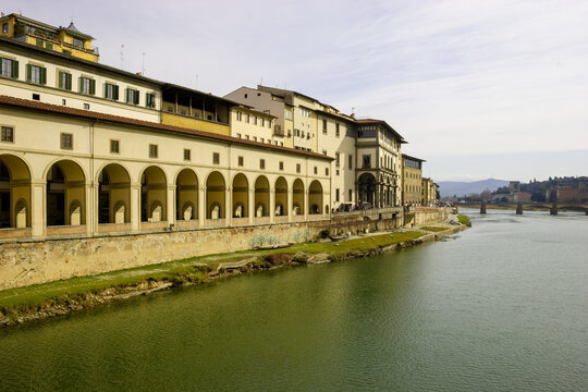 The Arno River flows past the Uffizi Gallery in Florence, Italy, with its distinctive arcades and historic architecture under a cloudy sky.