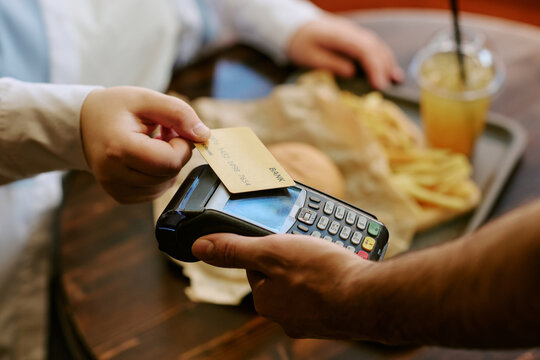 Caucasian young adult woman holding credit card making contactless payment on card reader while male hand assisting transaction, fast food meal with fries and drink visible in background