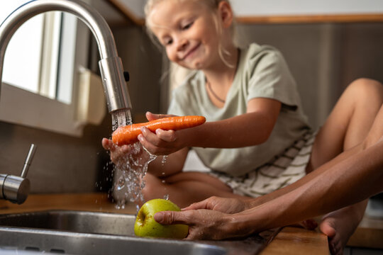 Little boy carefully washing a fresh carrot under running water while adult holds green apple in the kitchen sink