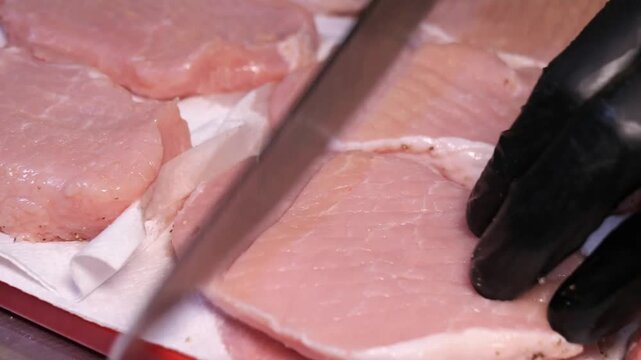 A chef scores and tenderizes raw pork chops with a knife and specialized tool on a cutting board in a professional kitchen.