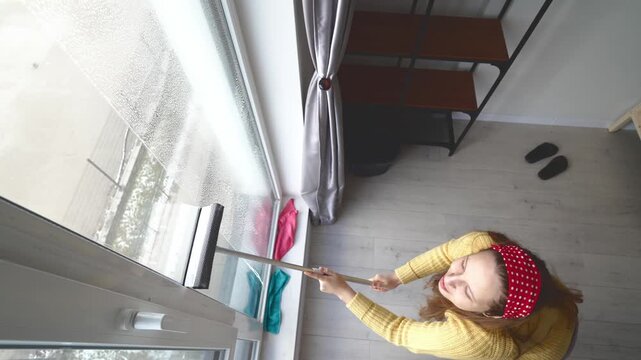 Woman Tidies Window Cheerfully. Joyful Lady With Yellow Sweater Polishes Glass Area Confidently