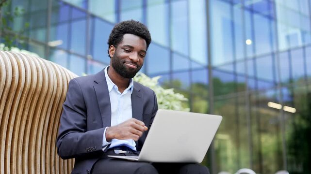 Satisfied happy african american businessman received great news on laptop sitting on a bench on street near a office building. Smiling glad entrepreneur in formal suit reads a positive good message