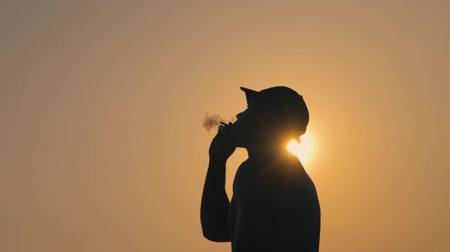 Silhouette of a man smoking during a golden sunset with the sun peeking behind him. Beautiful backlight and slow-motion create a relaxing and cinematic moment