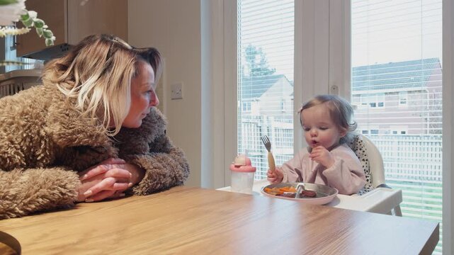 Grandmother babysitting her granddaughter as the toddler eats dinner at home, showing family care, bonding, childcare and a warm domestic mealtime scene indoors