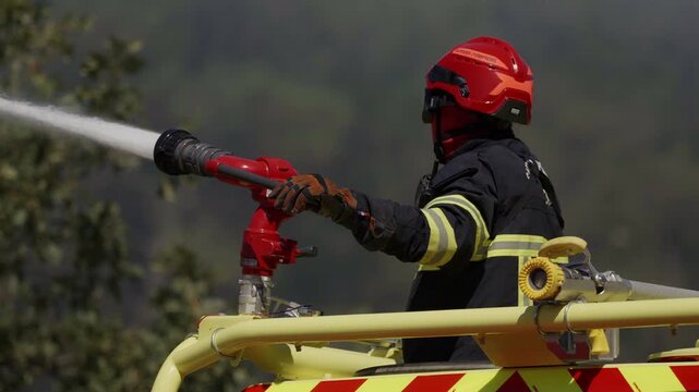 Firefighter in full protective gear and red helmet aims a high pressure water cannon mounted on a fire truck surrounded by green trees during an active intervention