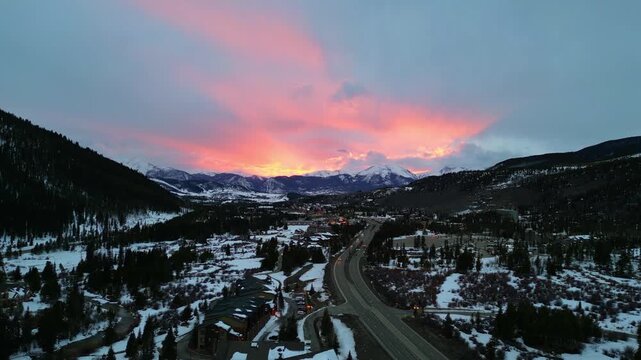 A stunning aerial view of a snowy mountain valley at sunset with vibrant pink and orange clouds above a winding highway and small town. Keystone, Colorado