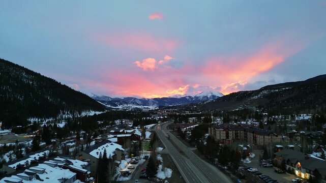 A breathtaking aerial shot of a snowy mountain town at sunset, featuring vibrant pink and orange clouds above rugged peaks and pine forests. Keystone, Colorado.