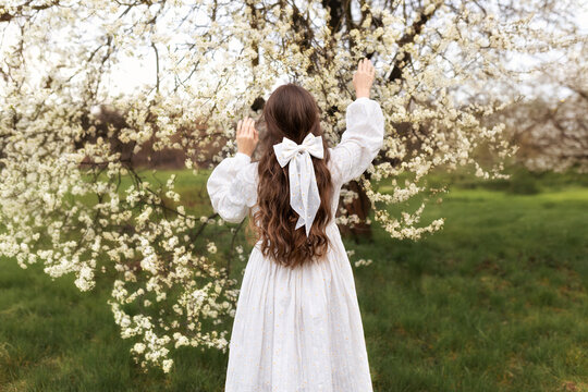 Back view of a young woman with long hair and a white bow, standing in a blooming orchard and reaching toward delicate spring blossoms. Concept of spring, beauty, femininity, and harmony with nature.