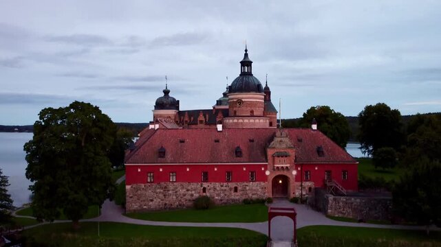 Gripsholm Castle in Mariefred, Sweden, is depicted with its distinct red-brick walls and round towers. The castle, surrounded by lush greenery, stands prominently near the water's edge of Lake 