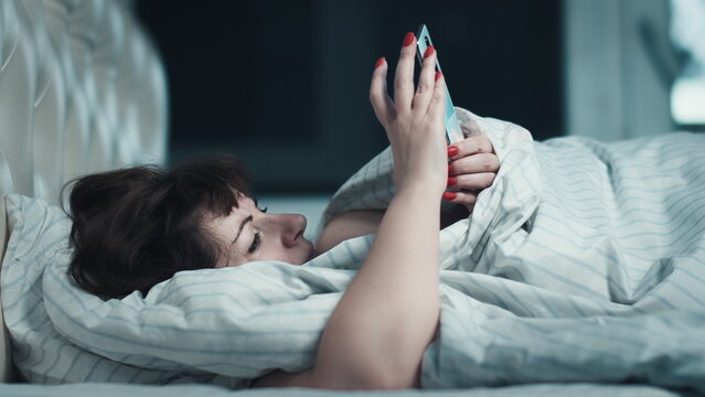 sleepy middle-aged woman lies on the bed and uses the phone at night, have fun and enjoy