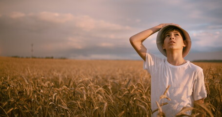 Obraz premium funny boy in a hat golden wheat field looks at the beautiful sky at sunset before storm, have fun and enjoy