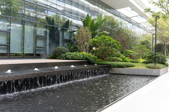 Modern urban water feature with cascading fountain and lush greenery in front of glass building,