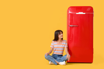 Beautiful young happy woman sitting near red fridge on yellow background