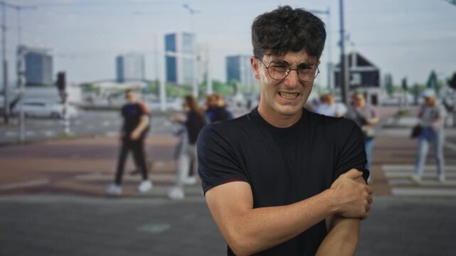 Man clutching his left arm and grimacing on a street crosswalk amid blurred pedestrians; discomfort vulnerability.
