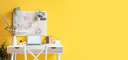 Workplace with laptop, blossoming tree branches and pegboard on yellow wall in room. Banner for...