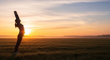 Fototapeta premium Silhouette of woman stretching arms up toward golden sunrise in wide open field with mist and morning light representing health and freedom concept