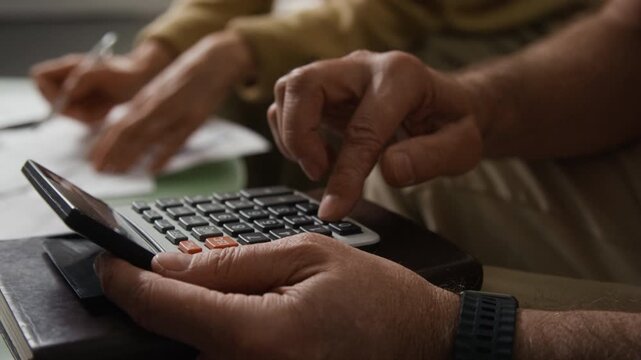 Extreme close-up shot of hands of anonymous senior man using calculator to count utility expenses