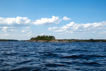 Naklejka premium Picturesque rocky island covered with green forest surrounded by the deep blue wavy waters of Lake Ladoga. Beautiful bright summer day with fluffy white clouds.