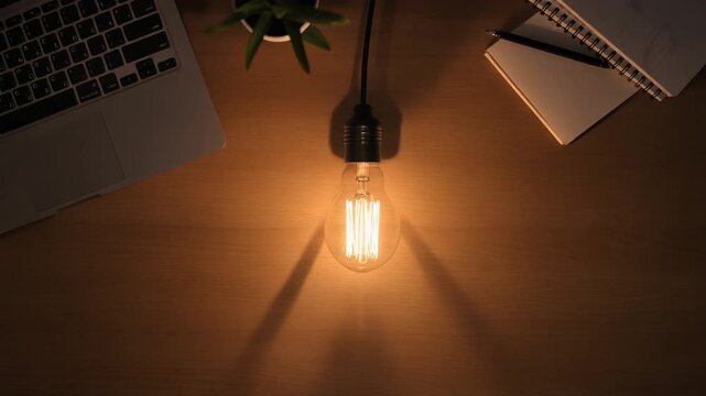 Overhead view of a desk with a glowing light bulb, laptop, plant, and notebook with a pen on a dark background.