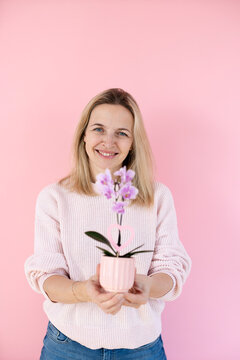 A blonde woman wearing a pink sweater is holding an orchid and standing in front of a pink background
