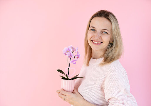 A blonde woman wearing a pink sweater is holding an orchid and standing in front of a pink background