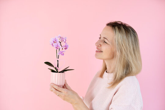 A blonde woman wearing a pink sweater is holding an orchid and standing in front of a pink background