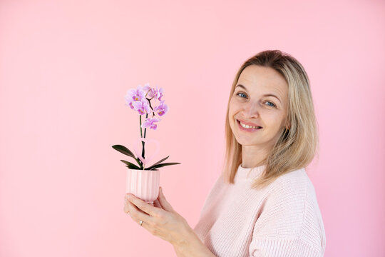 A blonde woman wearing a pink sweater is holding an orchid and standing in front of a pink background