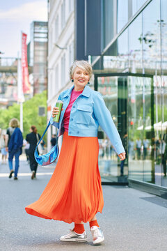Stylish woman in her fifties with grey hair walking in modern city street, confident mature female in bright outfit holding coffee, active urban lifestyle and fashion portrait outdoors