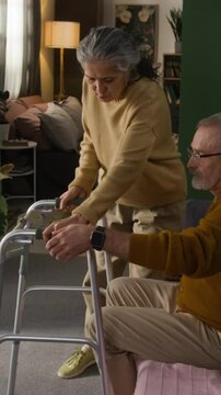 Vertical view of senior woman bringing walking frame for injured man and helping him stand up from couch in home setting