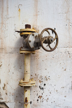 Old industrial valve with metal handwheel mounted on vertical pipeline near concrete wall. Heavy corrosion and rust indicate abandoned equipment and long-term neglect in industrial environment