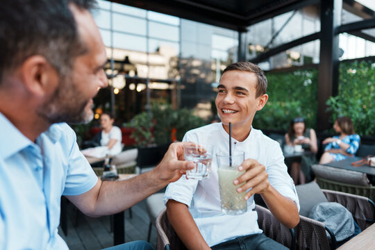 A happy father and his teenage son enjoy a refreshing drink while bonding at an outdoor cafe terrace during a summer afternoon.
