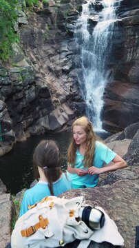 Happy mother and daughter talking by a forest waterfall in Norway. Bright northern daylight, authentic family bonding and sustainable travel concept.