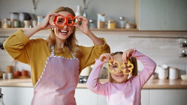 Funny mom child holding pepper slices peeping through at bright kitchen portrait
