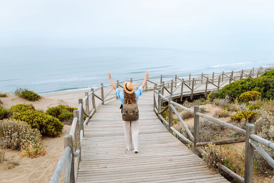 A cheerful woman wearing a hat and carrying a backpack walks along a wooden walking path overlooking the sea. A beautiful hiker enjoys the seascape at sunrise. Concepts include relaxation, adventure, 