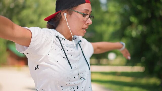 closeup expressive upperbody dance moves in park, young performer in backward cap and glasses practicing choreography with earphones, energetic hand gestures, candid rehearsal shots, summer light