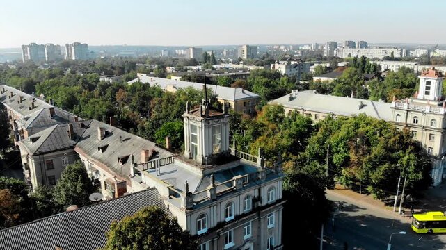 Aerial drone view of Sumy city center in Ukraine featuring historic architecture with a classical building topped by a Ukrainian flag, surrounded by green trees and urban streets.