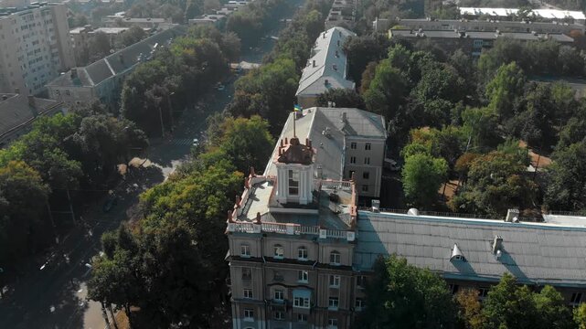 Aerial drone view of Sumy city center in Ukraine featuring historic architecture with a classical building topped by a Ukrainian flag, surrounded by green trees and urban streets.