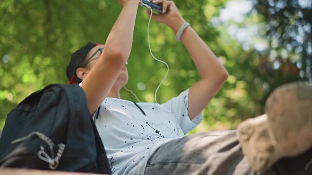 teen playing handheld console on bench, glassesclad youth in backward cap scrolls through game with focused expression, backpack at side, dappled sunlight through trees, thumbs tapping controls,