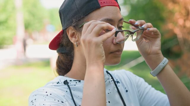 teen putting on glasses at park wearing backward cap and speckled hoodie, steady gaze and subtle smile, adjusting frames while scanning surroundings, warm summer light and shallow depth of field