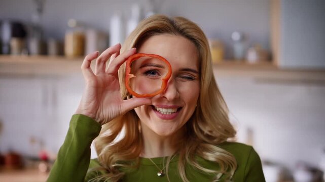 Funny woman holding pepper slice peeping through at bright kitchen portrait