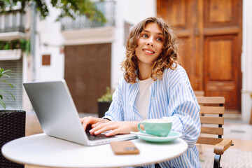 Fototapeta premium Portrait of female freelancer working on cafe terrace with cup of hot coffee. Beautiful woman sits with laptop outdoors in coffee shop. Concept of remote work, leisure, and business.