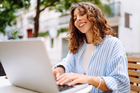 Portrait of female freelancer working on cafe terrace with cup of hot coffee. Beautiful woman sits with laptop outdoors in coffee shop. Concept of remote work, leisure, and business.