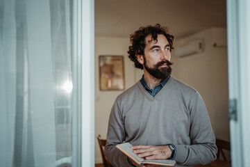 Fototapeta premium Pensive bearded man holding book looking away indoors
