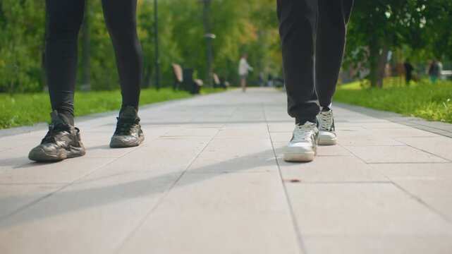 closeup feet walking on paved park path, two friends in sneakers and black pants pass wooden benches under leafy trees, warm afternoon light, relaxed stride and rhythmic footsteps, shallow cinematic