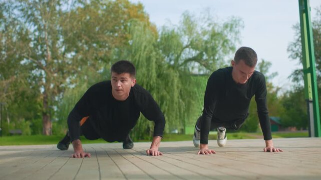 two men doing pushups outdoors on wooden deck in green park, training partners pushing through repetitions with focused expressions, athletic clothing, supportive teamwork and steady breathing
