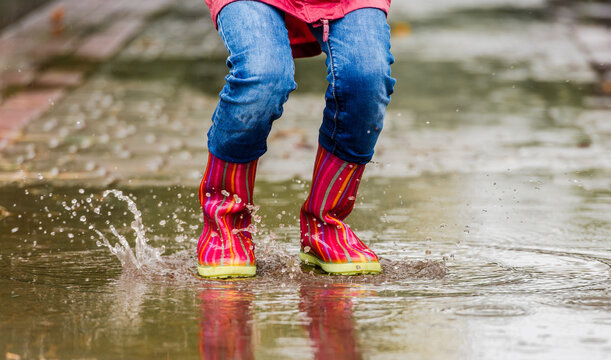 Child's Legs In Rubber Boots Splash Water While Jumping In A Puddle