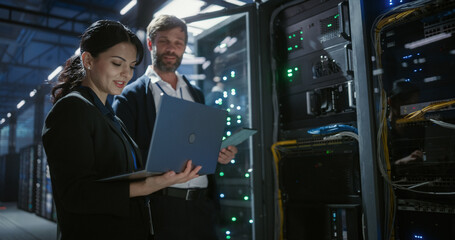 Two IT specialists use a laptop and tablet to inspect and analyze server racks in  data center,...