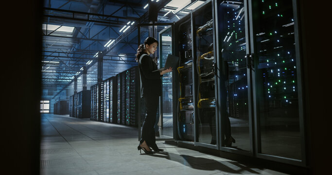 Technician uses a laptop computer to check and monitor server racks in a large data center, hardware installation, network devices diagnosttics, system check. Server room with mainframes.